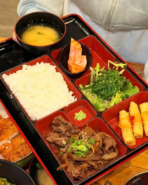 A red lacquer bento box with rice, teriyaki wagyu beef, spring rolls, salmon sashimi, salad and miso soup