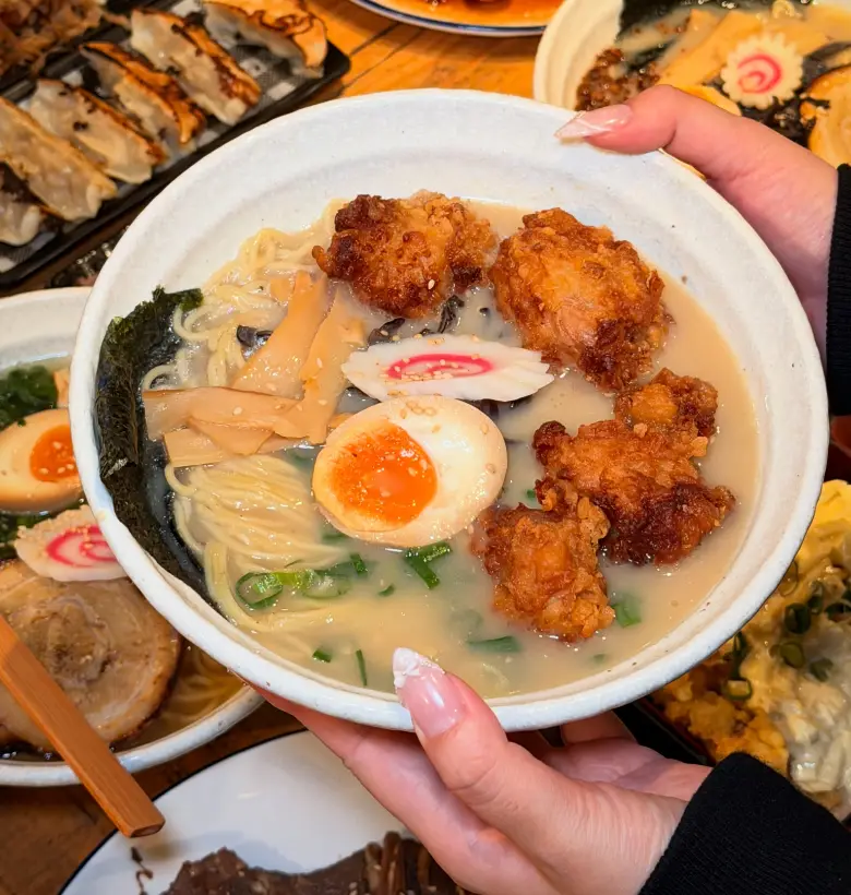 A bowl of tonkotsu ramen held up in two hands, topped with crispy karaage chicken, a jammy egg, menma, narutomaki and nori, with gyoza and more ramen on the table behind
